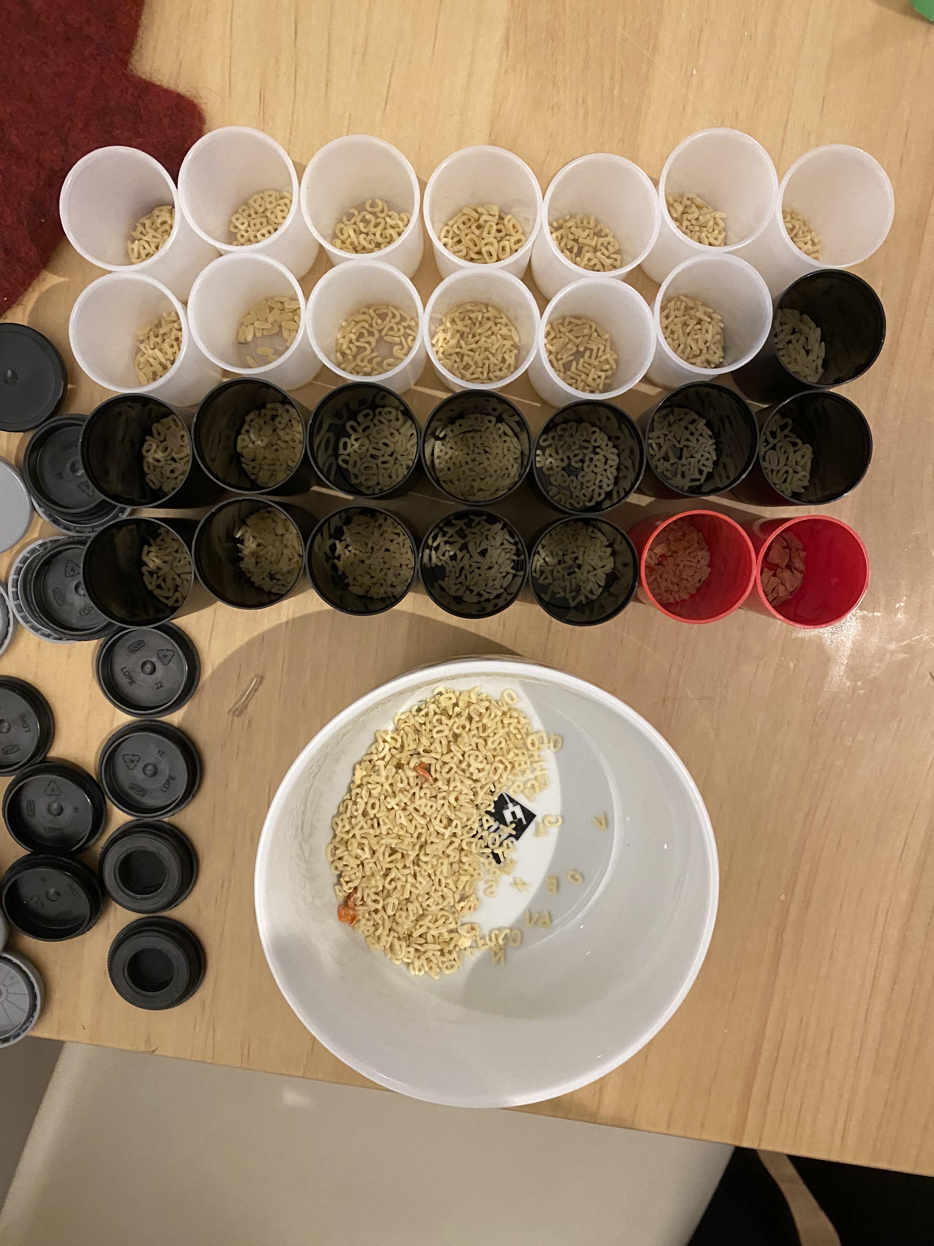 A grid of film canisters, each with one type of letternoodle and a bowl with more noodle in front.