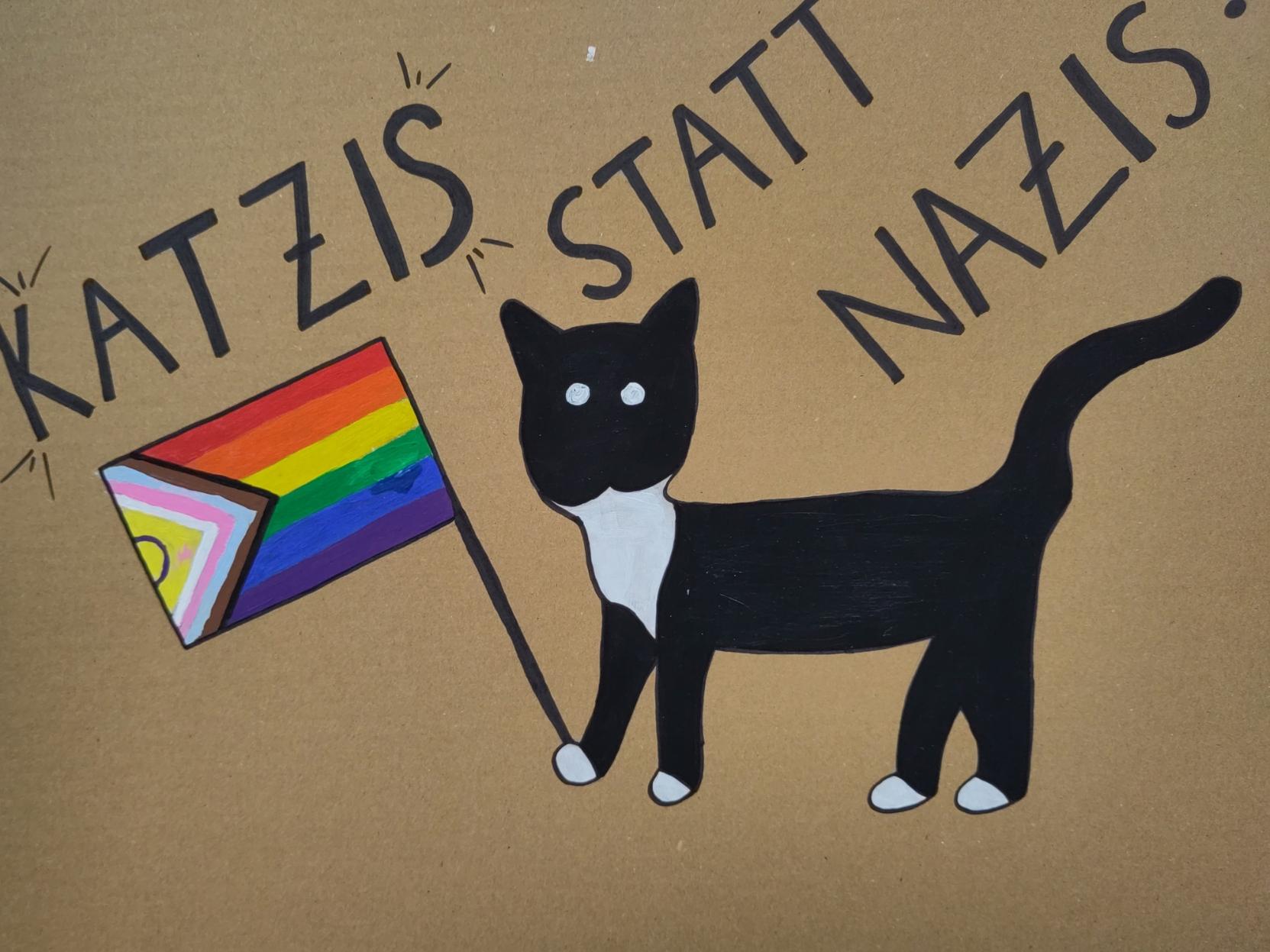 "Katzis statt Nazis" auf einem Pappschild. Darunter eine schwarz-weiß gemusterte Katze (aber hauptsächlich schwarz mit einer Progress-Pride-Flagge.